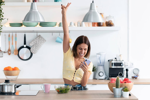 Фотография успешной молодой женщины, использующей свой мобильный телефон и празднующей что-то дома на кухне. - Фото, изображение