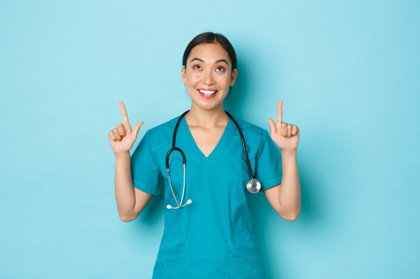 Covid-19, social distancing and coronavirus pandemic concept. Cheerful attractive asian female doctor, physician in scrubs pointing fingers and looking up with pleased happy expression - Photo, Image