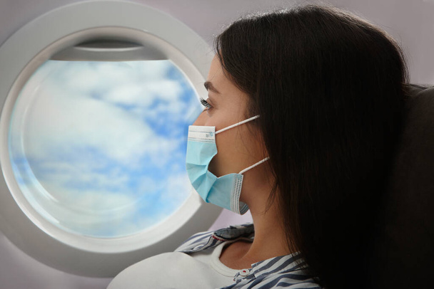 Traveling by airplane during coronavirus pandemic. Woman with face mask near porthole - 写真・画像