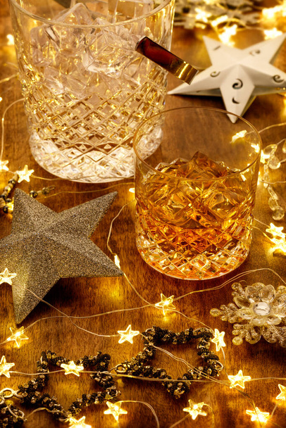 Κρύσταλλο ποτήρι ουίσκι και μια καράφα ουίσκι με χριστουγεννιάτικα φώτα, μπιχλιμπίδια και ασημένια αστέρια, σε ξύλινο τραπέζι - Φωτογραφία, εικόνα