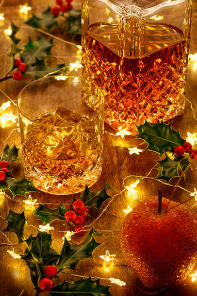 Carafe de whisky en cristal et un verre de whisky avec glace, lumières de Noël et baies de houx, sur une table en bois - Photo, image