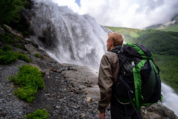 Man hiking with backpack looking at waterfall. Travel Lifestyle adventure concept, active vacations into the wild harmony with nature. North Caucasus, Dombai, Russia. - Photo, Image
