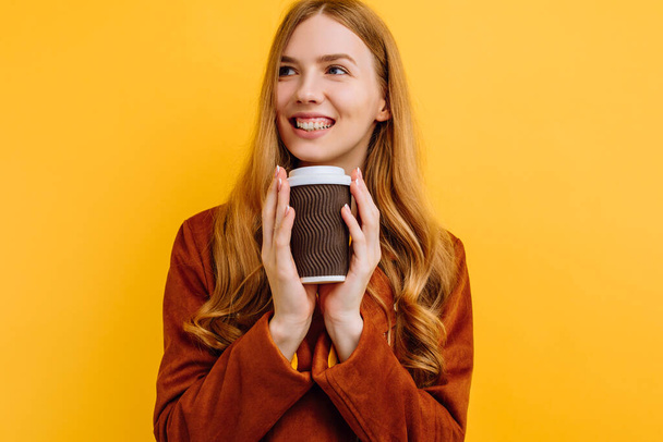 Pensive dreaming happy beautiful young woman in a yellow dress and autumn jacket, holding a paper Cup of coffee, on an isolated yellow background. The concept of autumn - Zdjęcie, obraz