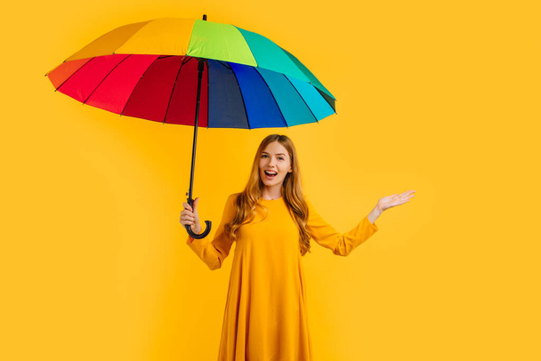 Happy attractive girl in a yellow dress posing with a rainbow umbrella, on an yellow background - Фото, изображение