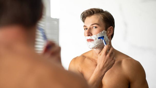 Jóképű fiatalember belenéz a tükörbe és borotválkozik. - Fotó, kép