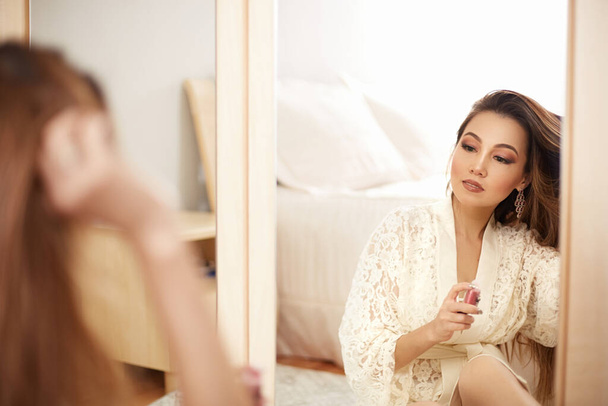 a young beautiful Asian woman in a beige lace dressing gown put on morning makeup and uses eye shadow, blush and Foundation . in the bedroom . shallow depth of focus. selective focus. - Фото, изображение