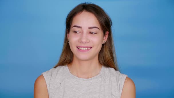 Gyönyörű vidám nő szürke pólóban körülnéz, mosolyog kék háttéren - Felvétel, videó