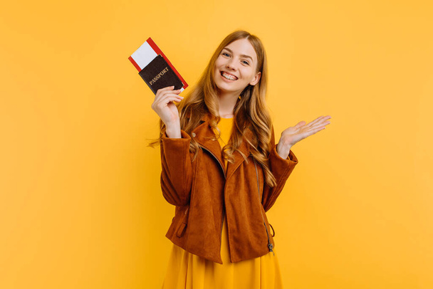 happy attractive girl in a yellow dress and autumn jacket, standing with her passport and tickets on an isolated yellow background - Foto, afbeelding
