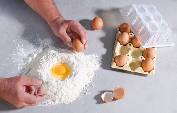 Mains masculines saisissant un œuf sur un tas de farine entouré de coquilles d'œufs et d'un carton d'œufs sur une table. Concept de cuisson. - Photo, image