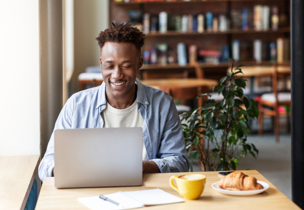 Счастливый чернокожий парень работает онлайн на ноутбуке в уютном городском кафе, свободное место - Фото, изображение