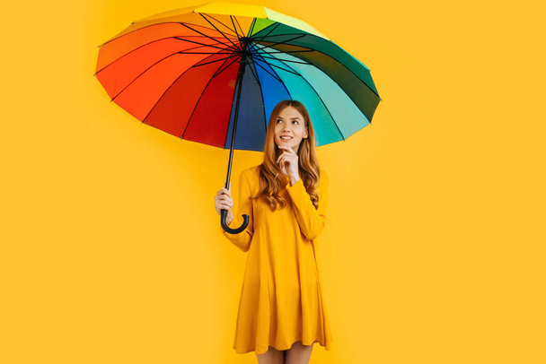 Pensive happy attractive girl, in a yellow dress, thinking and posing with a rainbow umbrella, on an yellow background - Photo, image