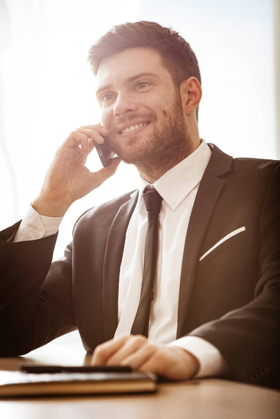 Bedrijfsconcept. Jonge zakenman zit aan de kantoortafel gelukkig te praten op een mobiele telefoon krijgt goed nieuws over zijn werk. Man in pak binnenshuis op glazen raamachtergrond. - Foto, afbeelding