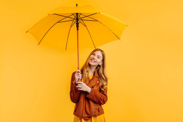 happy girl, in a bright yellow dress and an autumn jacket, stands with a yellow umbrella on an isolated yellow background - Photo, image