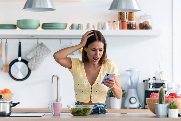 Tourné de belle jeune femme utilisant son téléphone portable tout en recevant de mauvaises nouvelles dans la cuisine à la maison. - Photo, image