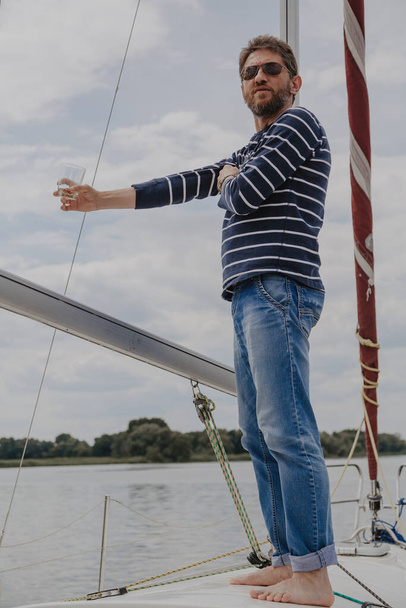 Businessman with a beard in a striped sweater stands on the nose of a white yacht and drinks whiskey from a glas - Photo, Image