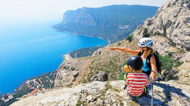 Child, young mother sit on mount top. Look at amazing sea landscape. Family travel adventure, hiking activity. Via ferrata tour with kids, exploring nature on summer vacation. Weekend day walking tour - Фото, зображення