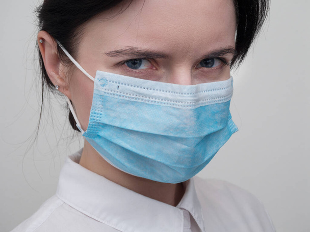 Portrait of a young European woman in a protective disposable medical mask. Concept of caronavirus Cavid 19 and human quarantine. Girl in a mask on a white wall. Masked medical worker - Photo, image