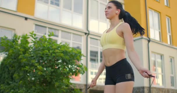 若い美しい女の子は、スポーツ服の遅い動きで公園のスキップロープにジャンプします。住宅を背景にスポーツをする - 映像、動画
