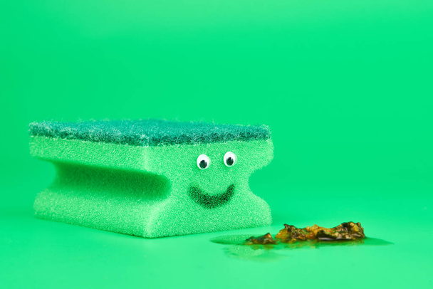 Vicces zöld szivacs szemekkel takarításhoz, takarításhoz és mosogatáshoz. Vicces ötlet. Puha és kemény kétoldalas szivacs. Minimális zöld háttér, másolás tér. - Fotó, kép