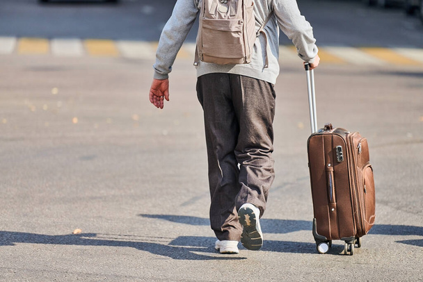 Stařík s kufrem na kolečkách. Muž s taškou na zavazadla kráčí po ulici od letiště. Návrat domů po výletě. Efekt časového posunu. Zadní pohled - Fotografie, Obrázek