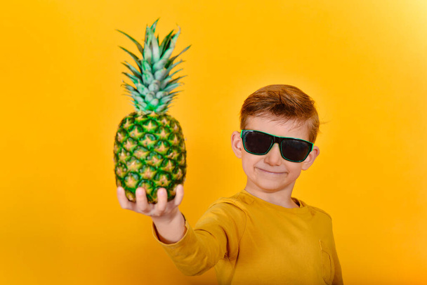 A boy in sunglasses holds a pineapple in his right hand. - Fotografie, Obrázek