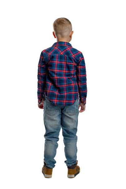 The boy is standing. A cute fat schoolboy in jeans, a shirt and a white T-shirt. Full height. White background. Vertical. Back view. - Foto, imagen