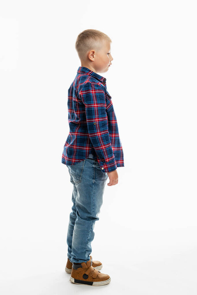 The boy is standing. A cute fat schoolboy in jeans, a shirt and a white T-shirt. Full height. White background. Vertical. Side view. - Foto, immagini