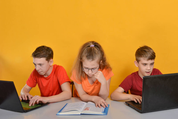 Τα παιδιά μαθαίνουν στο σπίτι, δύο αγόρια κάθονται σε ένα λάπτοπ, ένα κορίτσι διαβάζει βιβλία. Online μάθηση στο σπίτι έννοια. - Φωτογραφία, εικόνα
