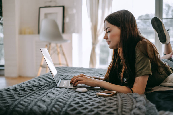 Attraktive junge kaukasische Frau, die mit einem Laptop arbeitet und zu Hause auf einem Bett liegt. Die Menschen arbeiten von zu Hause aus. Hochwertiges Foto - Foto, Bild