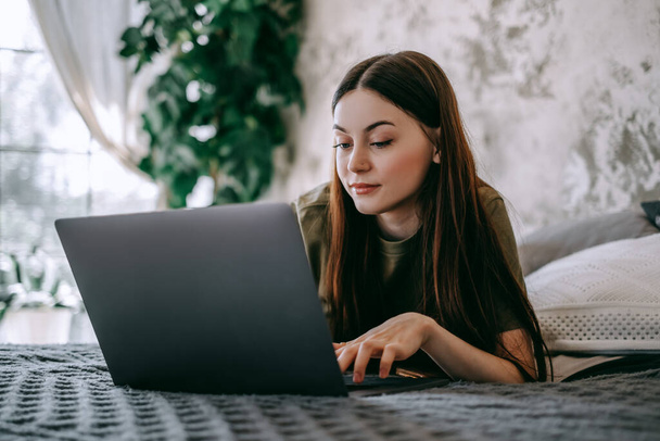 Attractive young caucasian woman working with laptop computer, lying on a bed at home. People work from home. High quality photo - Φωτογραφία, εικόνα