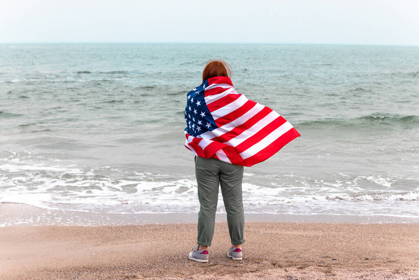 Kafkasyalı kızıl kadın Amerikan bayrağına sarılmış ve denize bakıyor. Bağımsızlık Günü için Amerikan bayrağı - Fotoğraf, Görsel