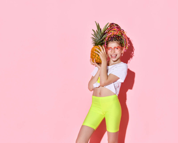 Teenage female with african pigtails, in sunglasses. She is holding pineapple and smiling, posing on pink background. Close up - Foto, Imagen