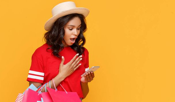 Elegante mujer étnica con bolsas de papel ofertas de descuento de navegación en el teléfono inteligente durante las compras contra el fondo amarillo - Foto, Imagen