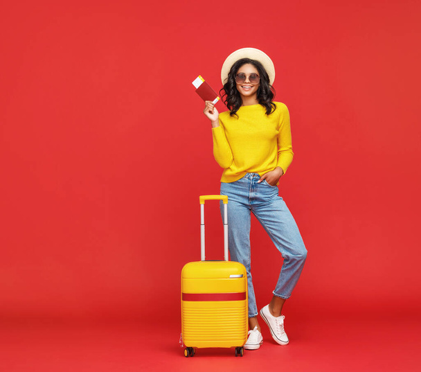 Mulher étnica de corpo inteiro na moda com passaporte e bilhetes sorrindo para câmera com bagagem durante as férias de verão contra backdro vermelho
 - Foto, Imagem