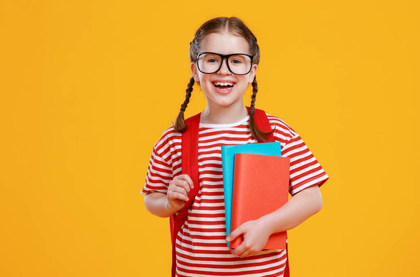 Χαρούμενο έξυπνο κορίτσι με σχολικά βιβλία να γελάει κατά τη διάρκεια των σχολικών σπουδών ενάντια στο κίτρινο backdro - Φωτογραφία, εικόνα