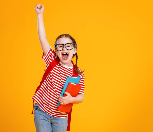 Podekscytowana dziewczyna w okularach i podręcznikach podnosząc rękę i krzycząc podczas świętowania sukcesu z żółtym backdro - Zdjęcie, obraz