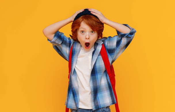 Astonished ginger boy with backpack holding hands on jowls head and looking at camera isolated on yellow backgroun - Fotoğraf, Görsel
