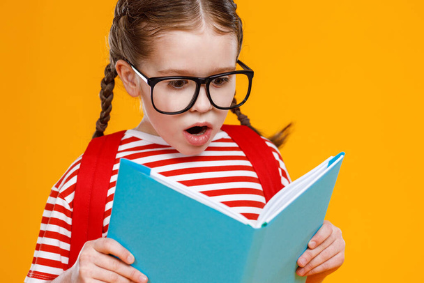 Elképesztő lány szemüvegben olvasás tankönyv nyitott szájjal, miközben tanul az iskolában ellen sárga backgroun - Fotó, kép