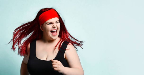 Close up portrait of young attractive plus-size model with flowing red hair, squinted eyes and naughty toothy smile on blue background. Copy space - Valokuva, kuva