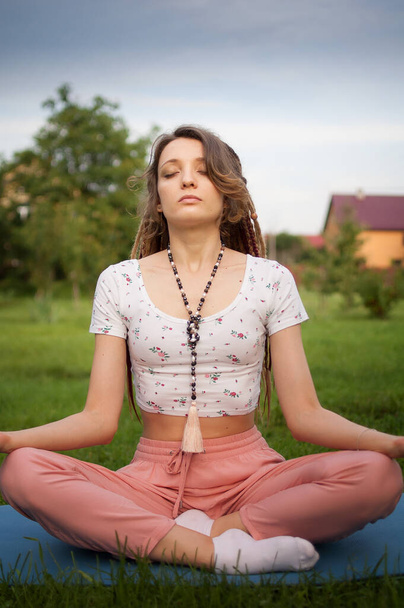 Young beautiful woman with long dreadlocks and beads made by natural stones is doing meditation and sitting in lotus position with closed eyes outdoors during morning on green grass of her backyard - Photo, Image