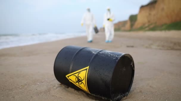 Dois cientistas de fatos de protecção brancos aproximam-se do barril negro com o sinal de perigo biológico que se encontra na praia.. - Filmagem, Vídeo