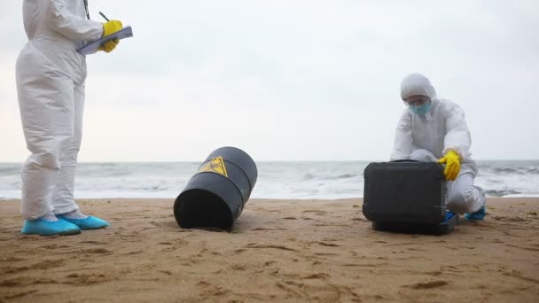 Naukowcy w białych kombinezonach ochronnych pracują w pobliżu niebezpiecznego obiektu na brzegu morza, zapisując dane na tablecie i przygotowując przyrządy do pobierania próbek. - Materiał filmowy, wideo