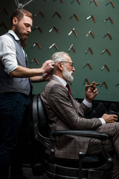 Bearded old businessman sitting at barber shop in chair choosing haircut design - Φωτογραφία, εικόνα