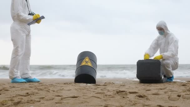 Deux scientifiques en combinaison de protection blanche recueillent les données et prennent des sondes du baril noir avec un signe de danger biologique sur le bord de la mer. - Séquence, vidéo