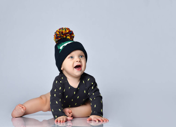 Little kid in black bodysuit and hat with croco print, barefoot. He is smiling, sitting on floor isolated on white. Close up - Foto, Bild