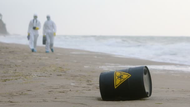 Dos hombres con trajes blancos de protección se acercan a un barril negro con un signo de peligro biológico, que yace en la orilla del mar. - Metraje, vídeo