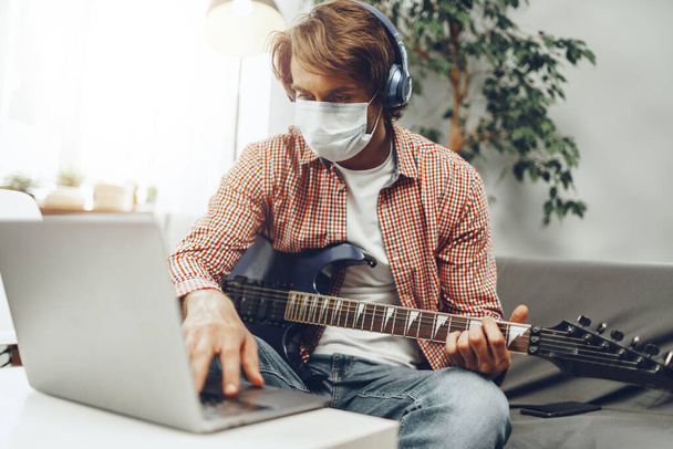 Young man plays guitar at home in medical mask. Coronavirus quarantine lifestyle concept - Photo, image