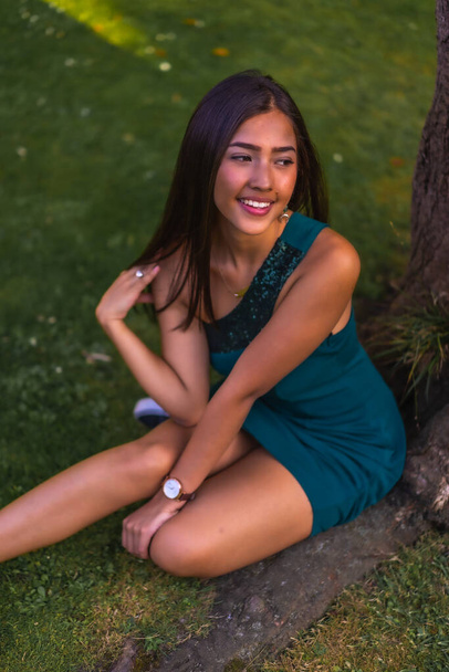 A young pretty brunette Latina with long straight hair leaning against a tree in a green dress. Portrait sitting on the grass in the park, shot flat - Fotografie, Obrázek