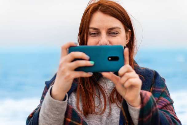 Kafkasyalı turist kızıl saçlı kadın gülümsüyor, fotoğraf çekiyor ya da sahilde, okyanus kenarında telefonla video kaydediyor. Tatil kavramı. - Fotoğraf, Görsel
