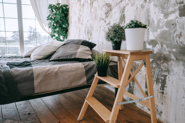 Bedroom with a large bed and wooden floor shelf with home plants in the sunny day, home comfort. interior details. High quality photo - Photo, image